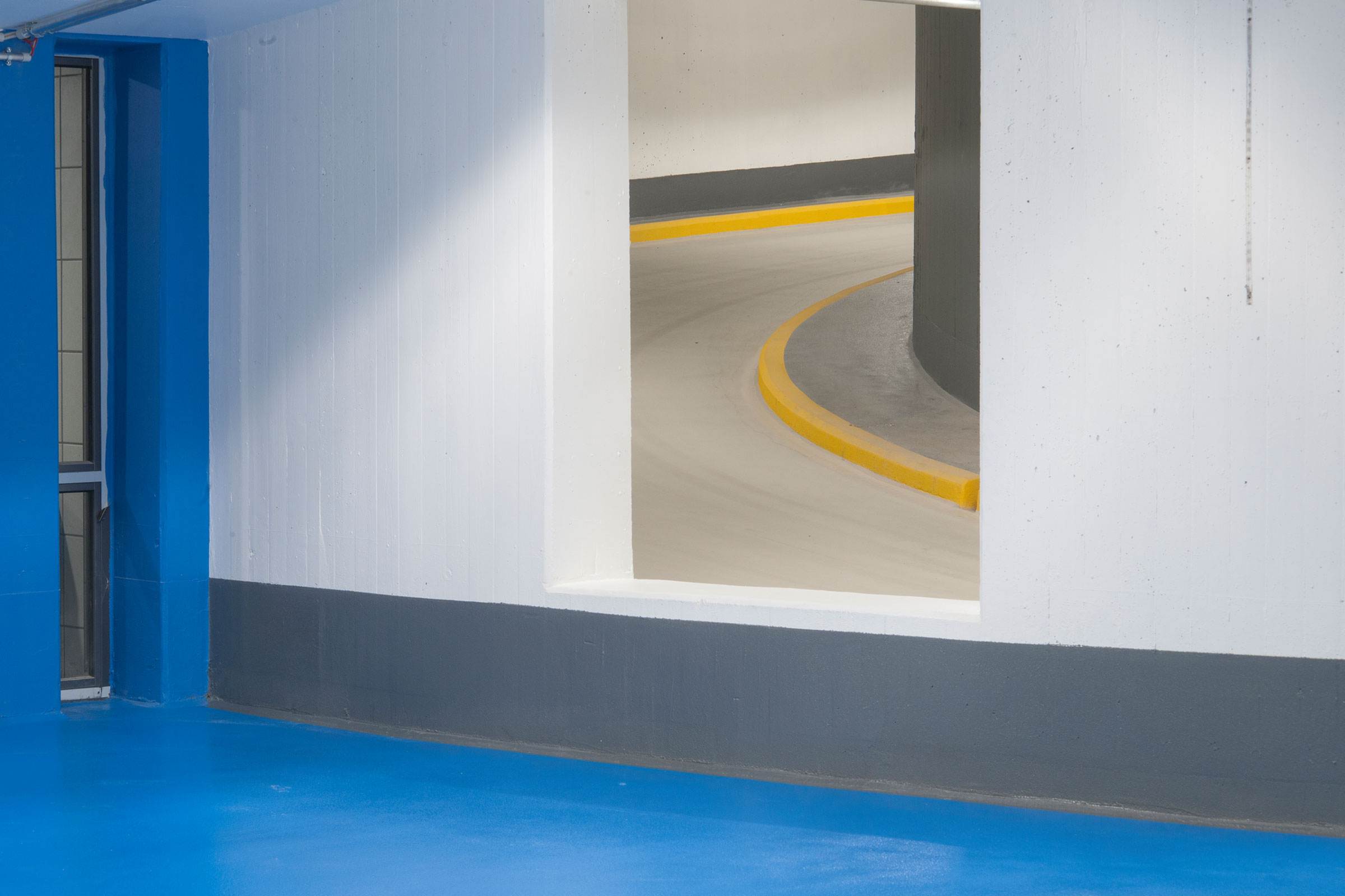 Vit väggyta i parkeringshus med blå gångbana intill.