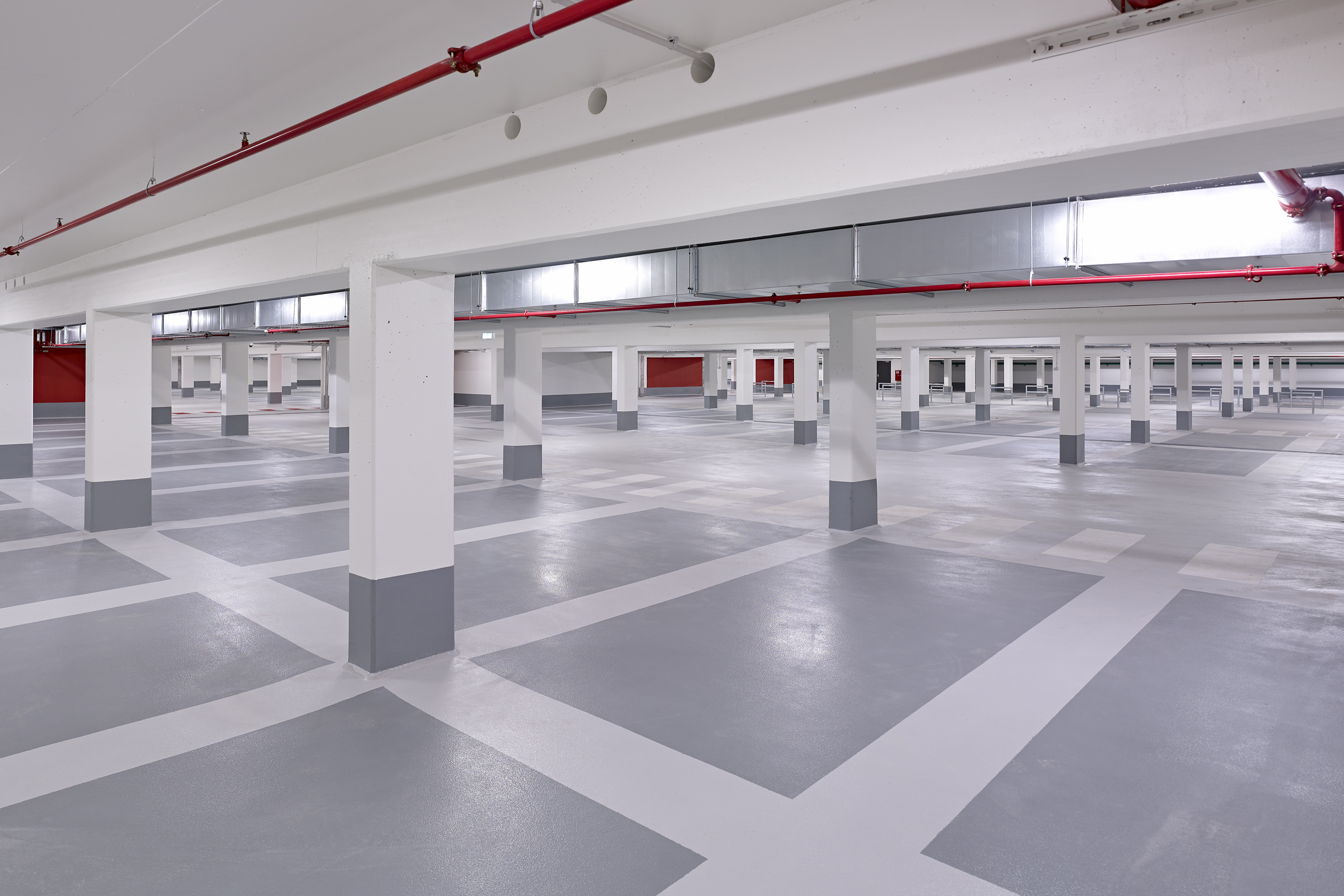 Stort utrymme i parkeringshus med flera gråa parkeringsfickor och vita pelare. Röda streck i taket.