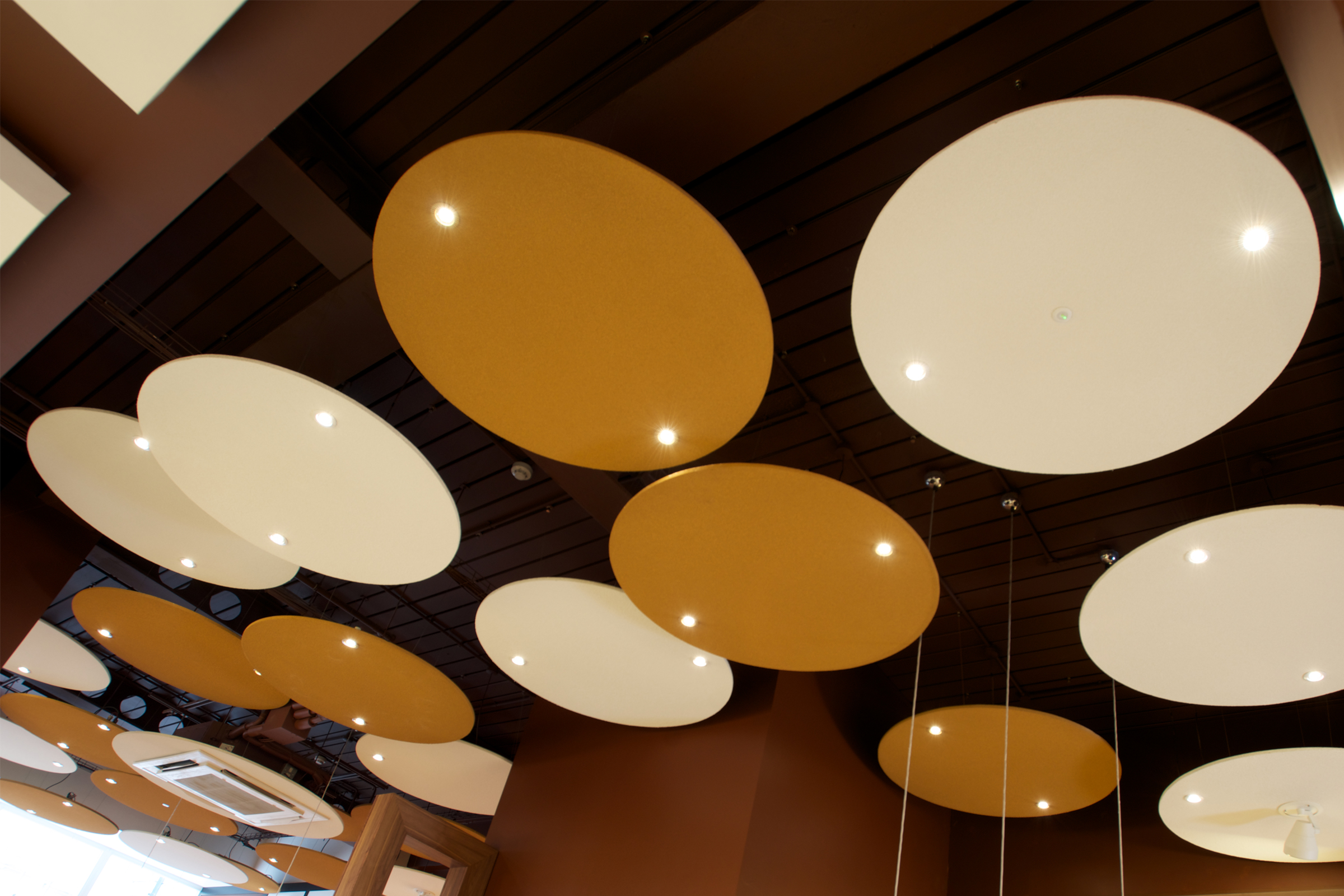 Runda akustikplattor i beiga och bruna kulörer med spotlights hänger ner från ett tak.